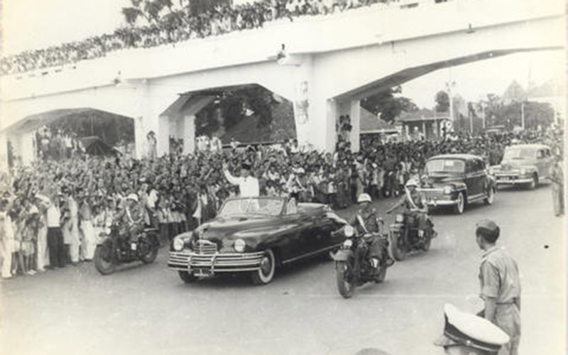 Presiden Sukarno berada di atas mobil terbuka yang sedang berjalan tengah melambaikan tangan membalas masyarakat yang berkerumun menyambutnya di pinggir jalan, diikuti dengan beberapa mobil kenegaraan lain. /Sistem Informasi Kearsipan Arsip Nasional Republik Indonesia